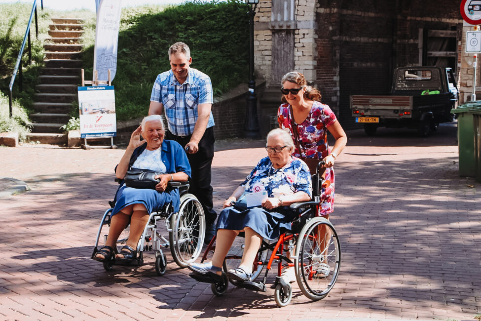 Twee oudere mensen in rolstoelen worden begeleid door een man en een vrouw op een zonnige dag buiten bij een stenen gebouw.