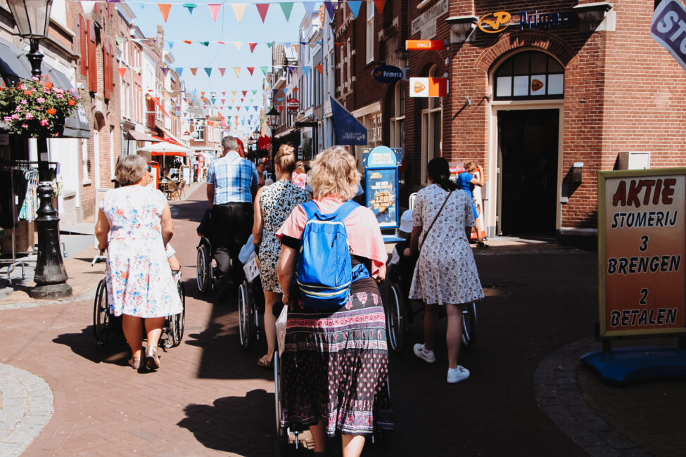 Groep mensen loopt door een zonnige winkelstraat met vlaggetjes boven hen. Enkelen duwen rolstoelen. Links een bloemenkraam.