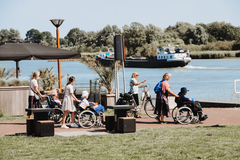 Mensen in rolstoelen genieten van een zonnige dag aan het water, met een passerend schip en groene bomen op de achtergrond.