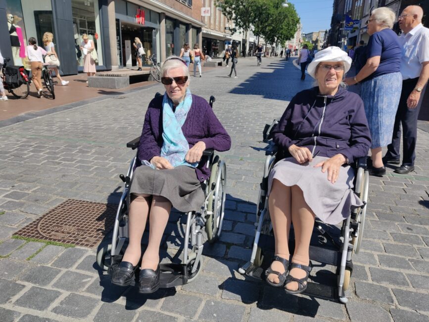 Twee oudere vrouwen in rolstoelen genieten van een zonnige dag op een winkelstraat, omringd door voorbijgangers en winkels.