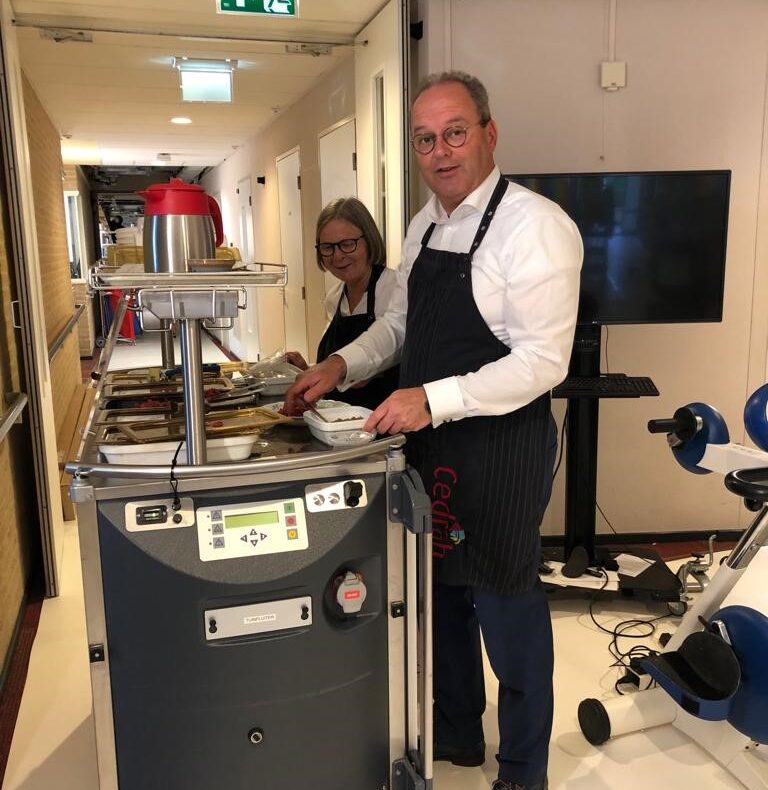 Twee mensen serveren maaltijden vanuit een mobiele kar in een gang. Ze dragen schorten en staan in een lichte ruimte.