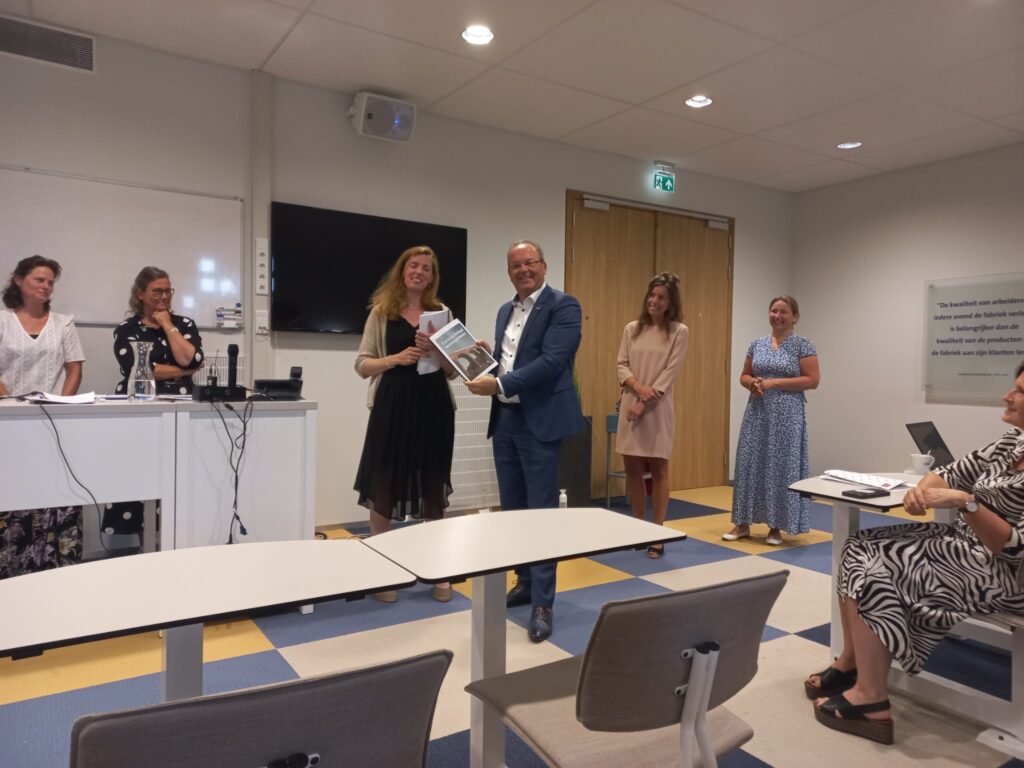 Groep mensen in een vergaderruimte. Een man en vrouw staan vooraan en houden een certificaat vast. Anderen kijken toe.