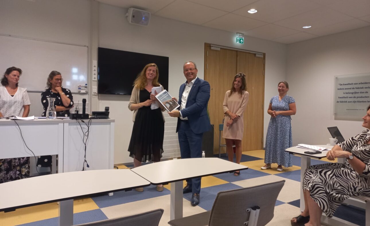 Groep mensen in een vergaderruimte. Een man en vrouw staan vooraan en houden een certificaat vast. Anderen kijken toe.