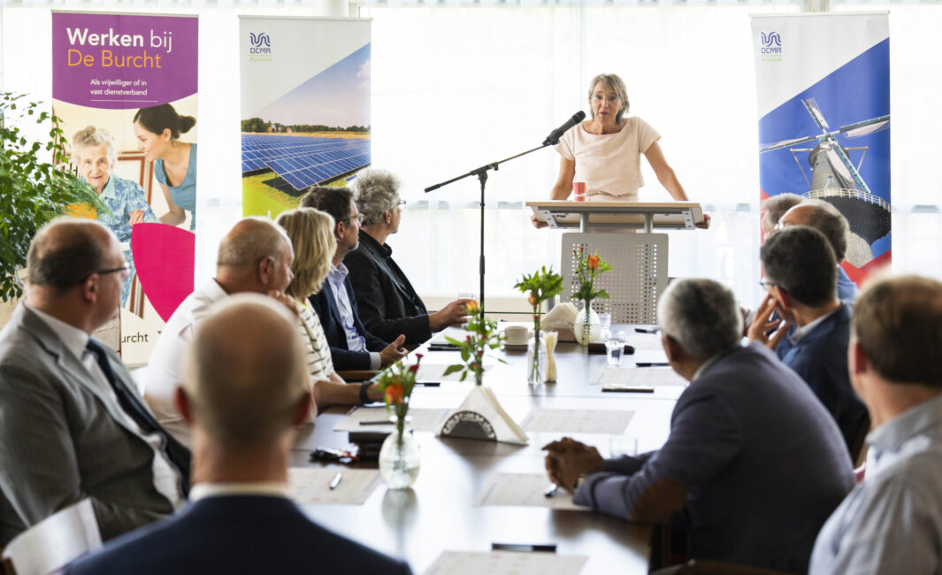 Een vrouw spreekt voor een groep mensen aan een tafel. Op de achtergrond zijn banners en bloemen op tafel te zien.