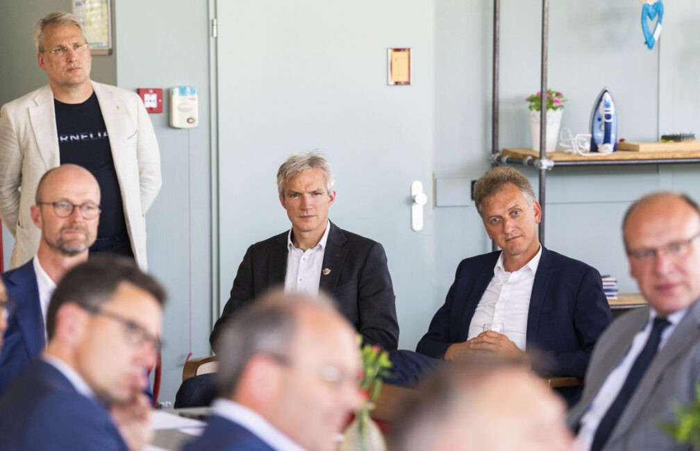 Groep mannen in pakken zit in een vergaderruimte. Ze luisteren aandachtig naar een spreker buiten beeld. Tafels met bloemen.