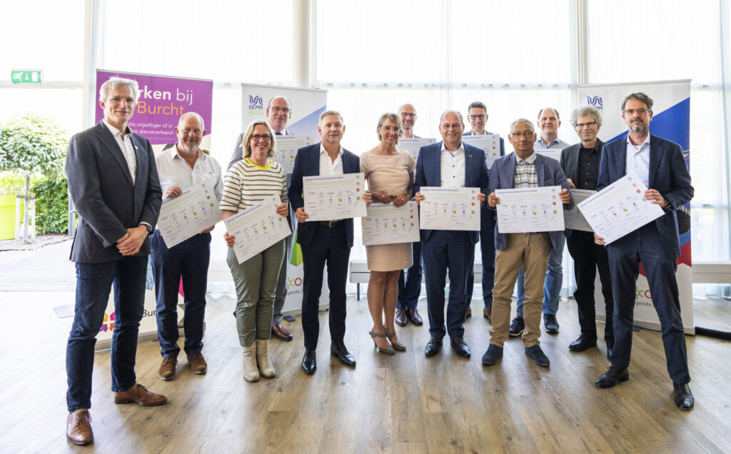 Groep mensen poseert binnen met certificaten in hun handen. Ze staan voor banners en kijken naar de camera.