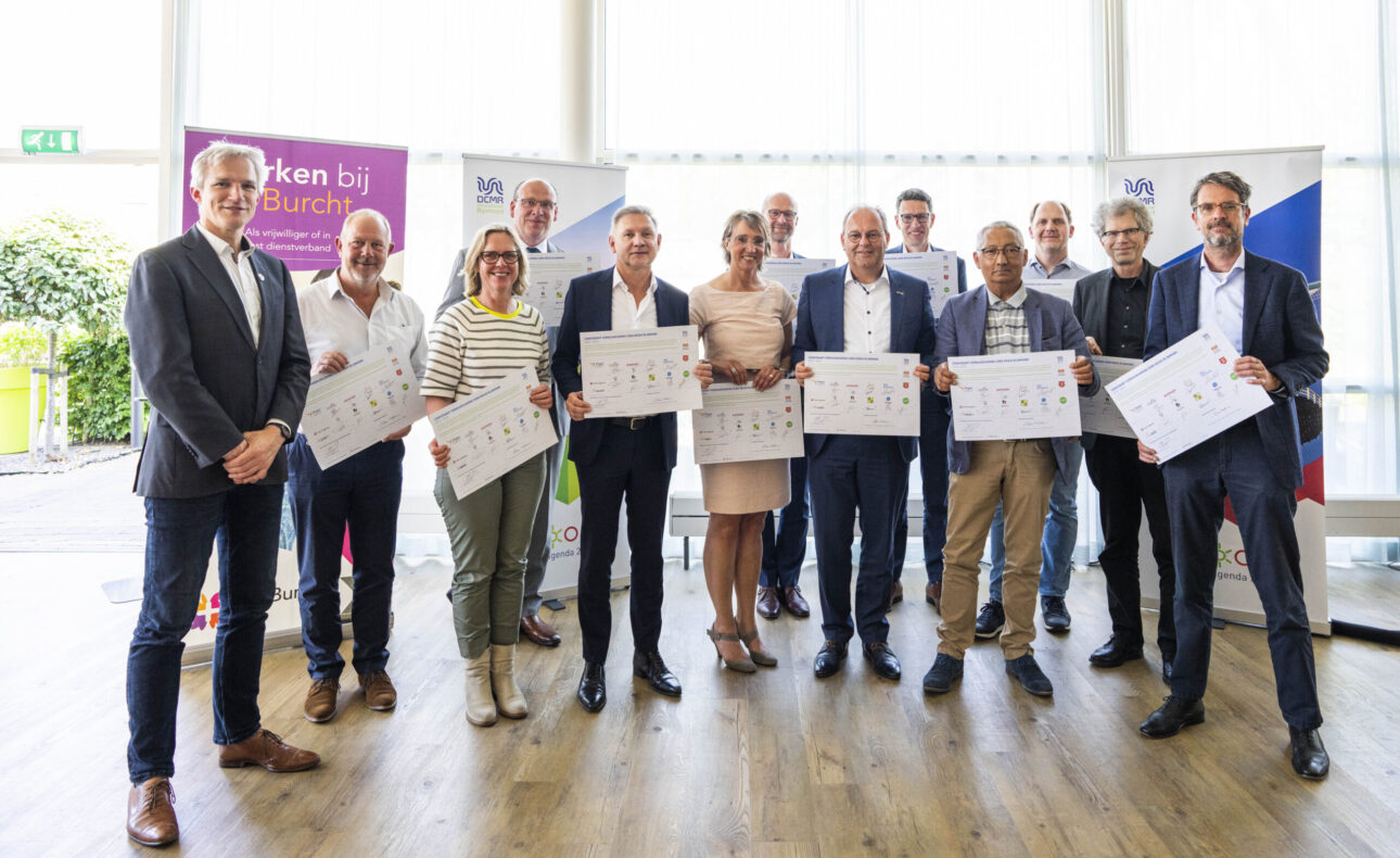Groep mensen poseert binnen met certificaten in hun handen. Ze staan voor banners en kijken naar de camera.
