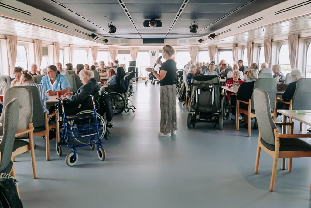 Een groep mensen zit in een ruime zaal met grote ramen. Er zijn rolstoelen en rollators zichtbaar. Een vrouw staat in het midden.