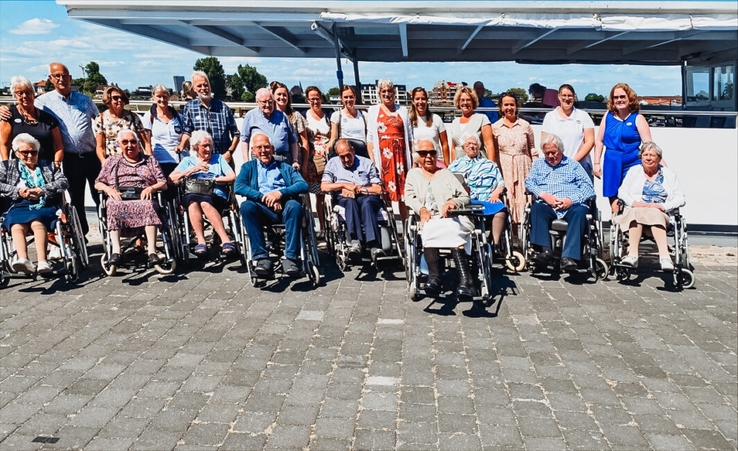 Groepsfoto van ouderen en verzorgers buiten op een zonnige dag. Sommigen zitten in rolstoelen, anderen staan erachter.