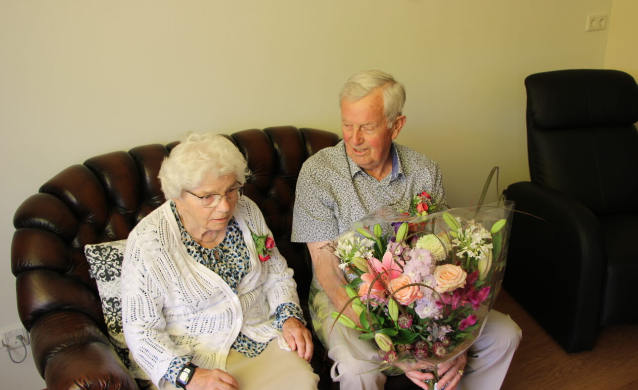 Oudere man en vrouw zitten op een bruine bank. De man houdt een grote bos bloemen vast. Ze kijken beiden naar voren.