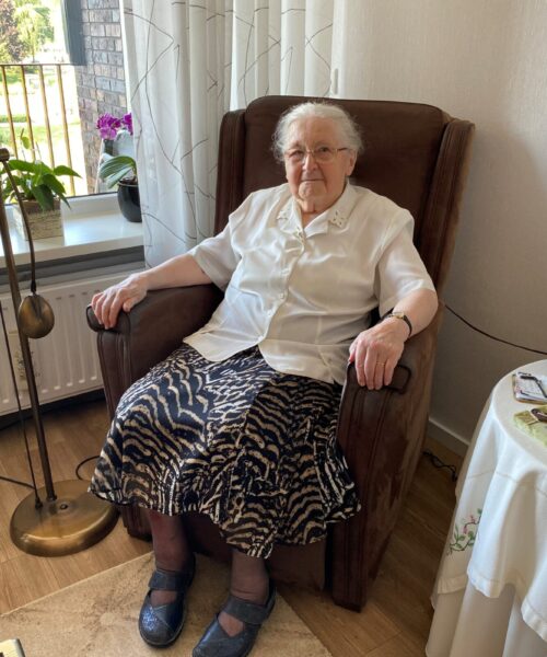 Oudere vrouw zit glimlachend in een bruine fauteuil in een gezellige woonkamer met planten en een tafellamp naast haar.