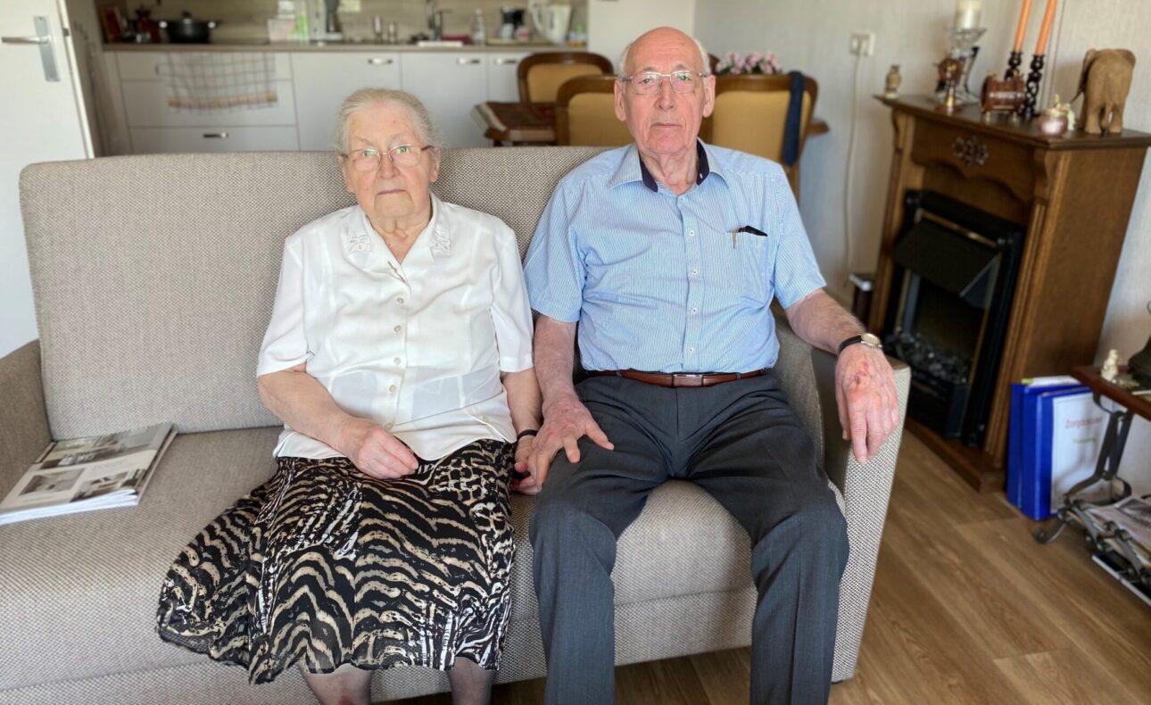 Ouder echtpaar zit samen op een bank in een woonkamer. De vrouw draagt een witte blouse en rok, de man een blauw overhemd.