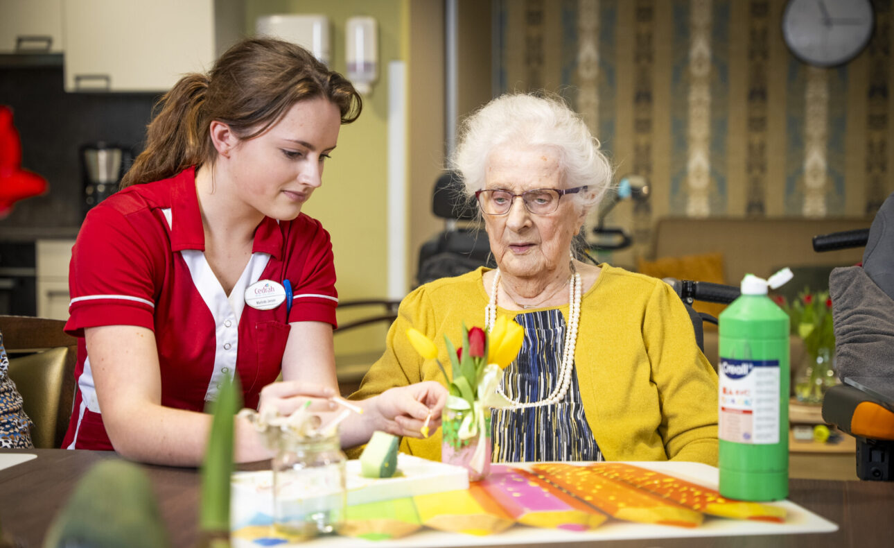 Een zorgmedewerker helpt een oudere vrouw met een creatieve activiteit aan tafel in een verzorgingshuis.