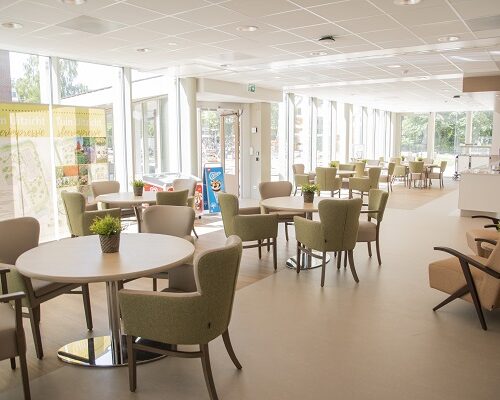 Een lichte, moderne gemeenschappelijke ruimte met tafels en stoelen, grote ramen en uitzicht op een groene omgeving.