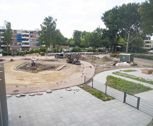 Bouwplaats met machines en materialen, omgeven door bomen en gebouwen, met een gedeeltelijk betegeld terrein op de voorgrond.