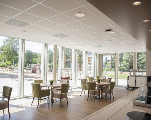Een lichte, moderne ruimte met grote ramen, tafels en stoelen. Buiten is groen te zien. Het is een uitnodigende omgeving.