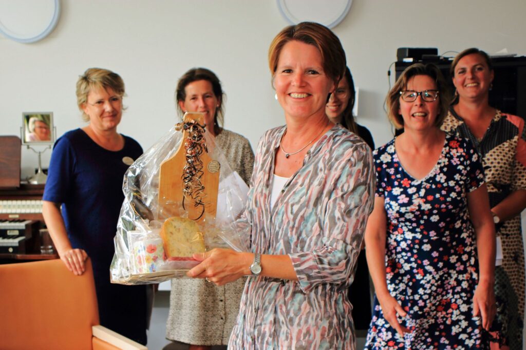 Vrouw in het midden houdt een cadeaupakket vast, omringd door lachende vrouwen in een kamer met een orgel op de achtergrond.