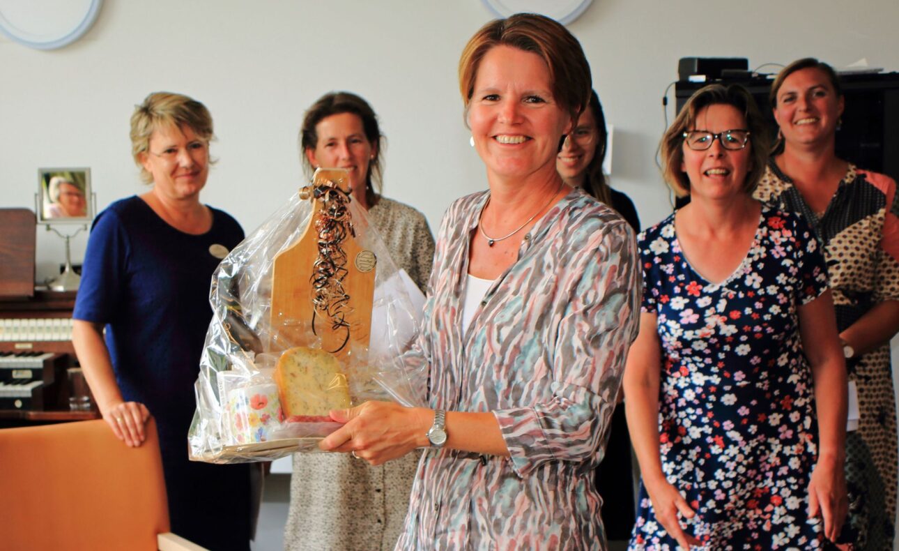 Vrouw in het midden houdt een cadeaupakket vast, omringd door lachende vrouwen in een kamer met een orgel op de achtergrond.