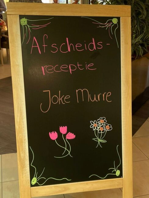 Een krijtbord met de tekst "Afscheidsreceptie Joke Murre" en tekeningen van bloemen in roze en wit op een zwarte achtergrond.