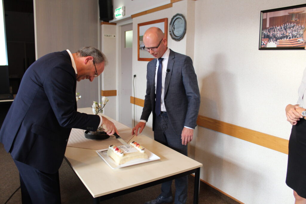 Twee mannen in pakken snijden samen een taart aan op een tafel in een vergaderruimte. Er hangt een schilderij aan de muur.