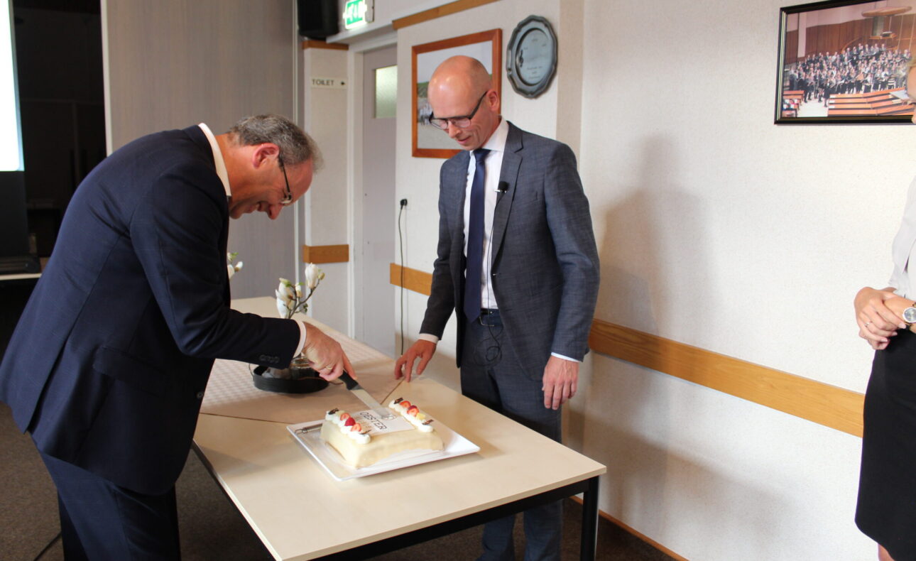 Twee mannen in pakken snijden samen een taart aan op een tafel in een vergaderruimte. Er hangt een schilderij aan de muur.
