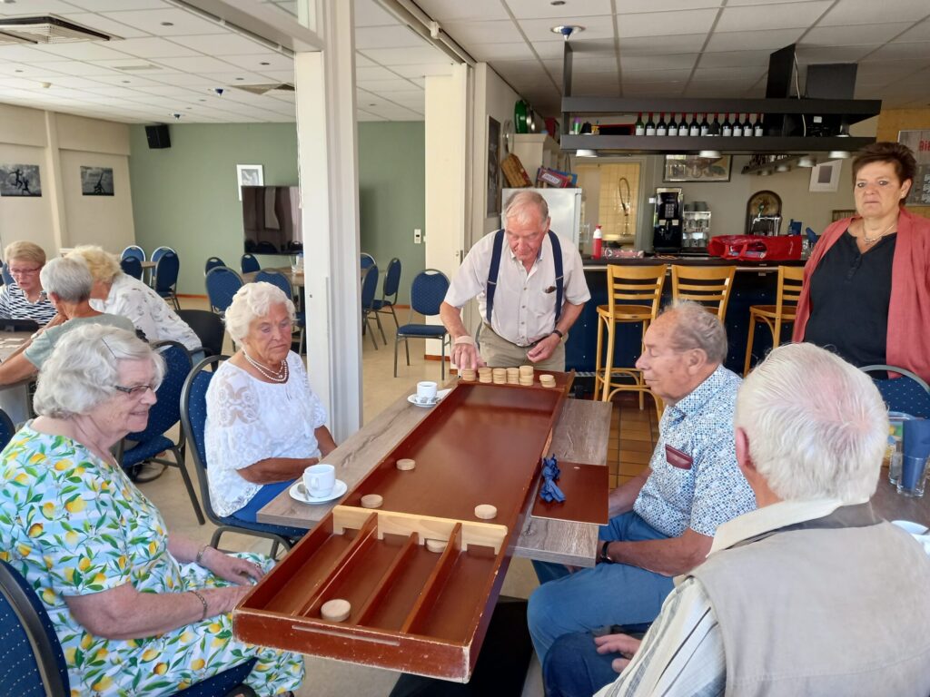 Een groep ouderen speelt sjoelen in een gemeenschappelijke ruimte. Ze zitten rond een tafel met koffie en thee.