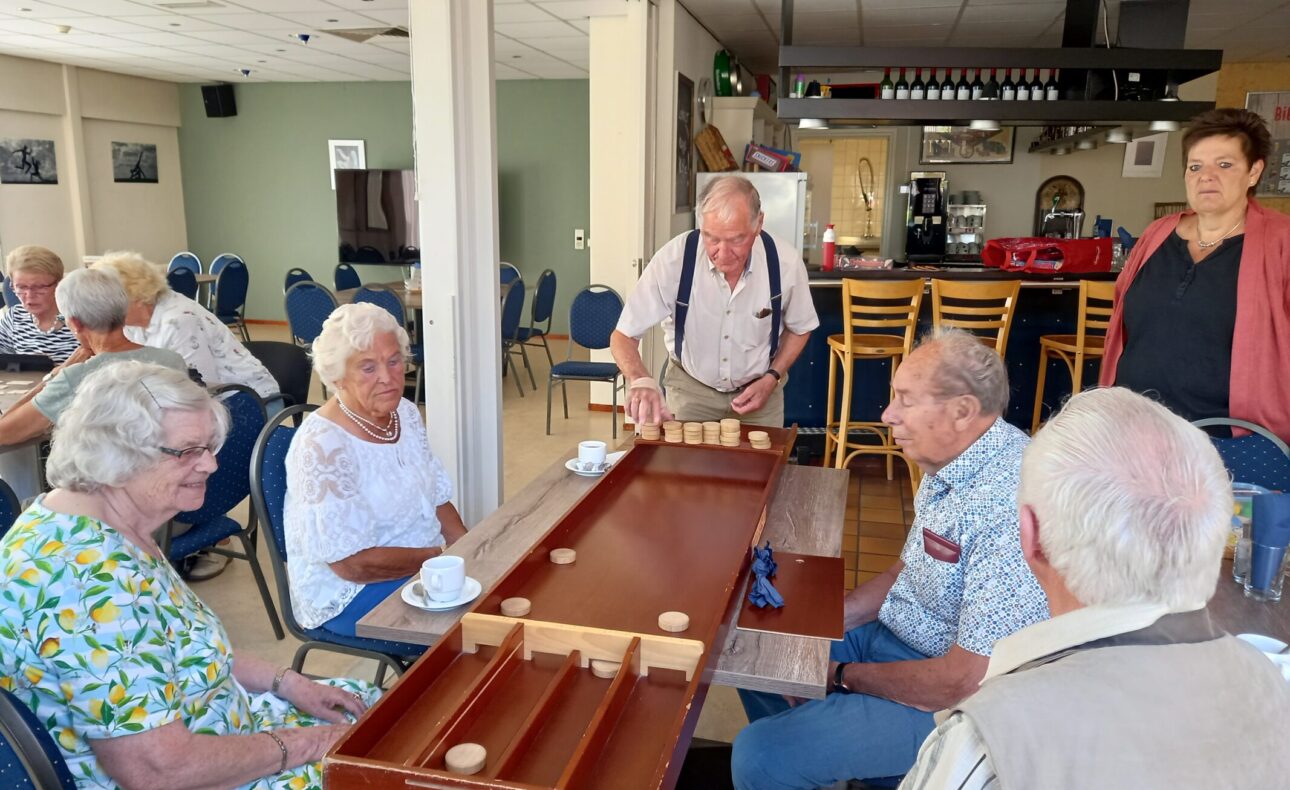 Een groep ouderen speelt sjoelen in een gemeenschappelijke ruimte. Ze zitten rond een tafel met koffie en thee.