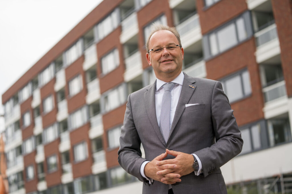 Man in grijs pak staat voor een modern gebouw met bakstenen en grote ramen. Hij glimlacht en heeft zijn handen gevouwen.