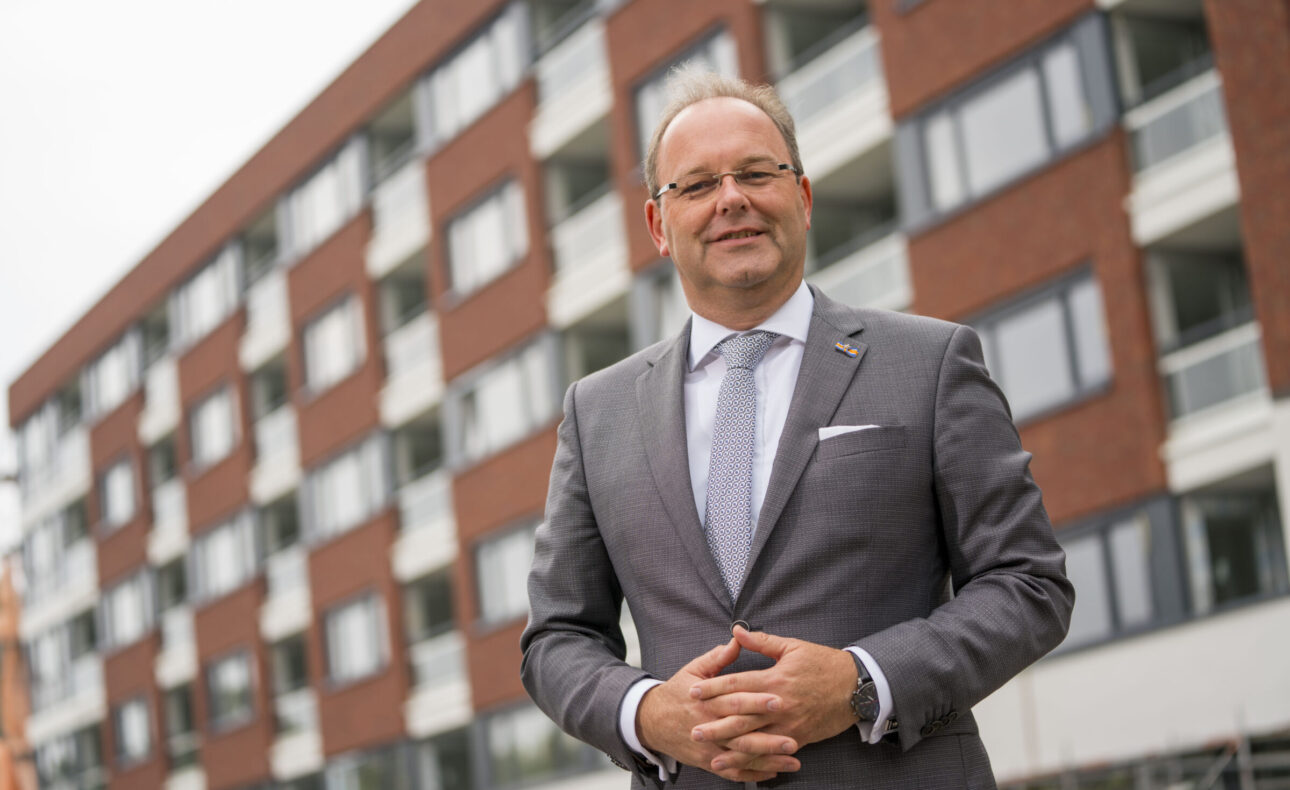 Man in grijs pak staat voor een modern gebouw met bakstenen en grote ramen. Hij glimlacht en heeft zijn handen gevouwen.