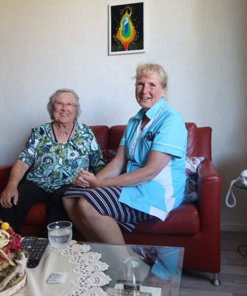 Twee vrouwen zitten glimlachend op een rode bank in een woonkamer. Er hangt een kleurrijk schilderij aan de muur.