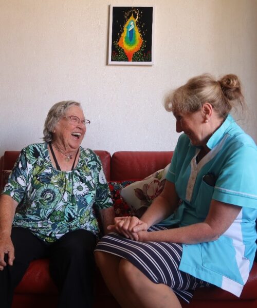 Twee vrouwen zitten lachend op een rode bank in een woonkamer. De vrouw links draagt een groene blouse, de vrouw rechts een uniform.