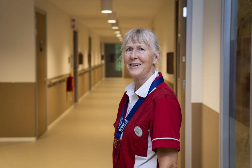 Een verpleegkundige in een rood uniform staat glimlachend in een goed verlichte gang van een zorginstelling, met deuren aan beide zijden.