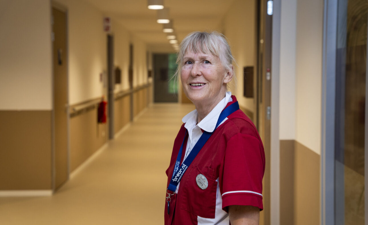 Een verpleegkundige in een rood uniform staat glimlachend in een goed verlichte gang van een zorginstelling, met deuren aan beide zijden.