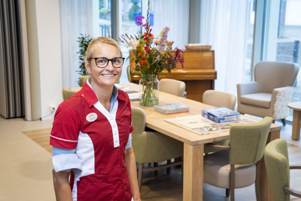 Vrouw in rood-wit uniform staat glimlachend in een lichte kamer met een houten tafel, bloemen en een piano op de achtergrond.