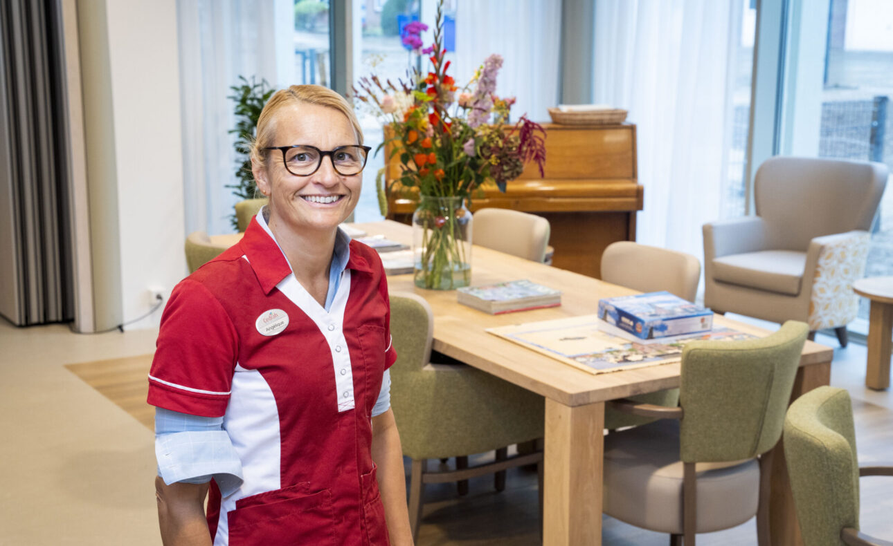 Vrouw in rood-wit uniform staat glimlachend in een lichte kamer met een houten tafel, bloemen en een piano op de achtergrond.