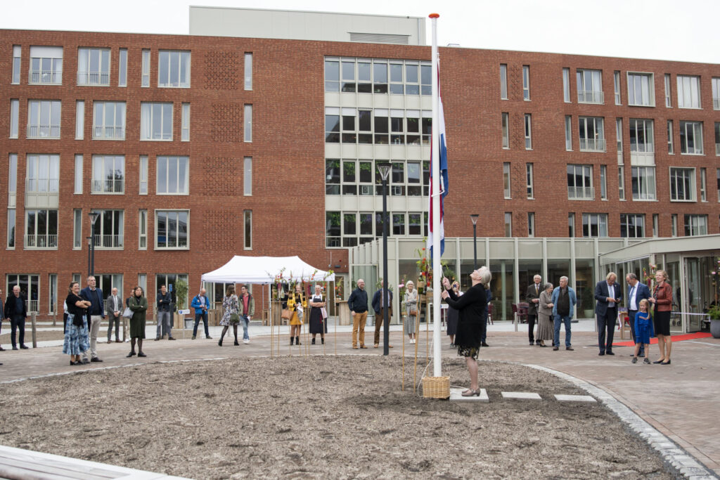 Groep mensen verzameld voor een modern gebouw met rode bakstenen, naast een vlaggenmast en een witte tent op de achtergrond.