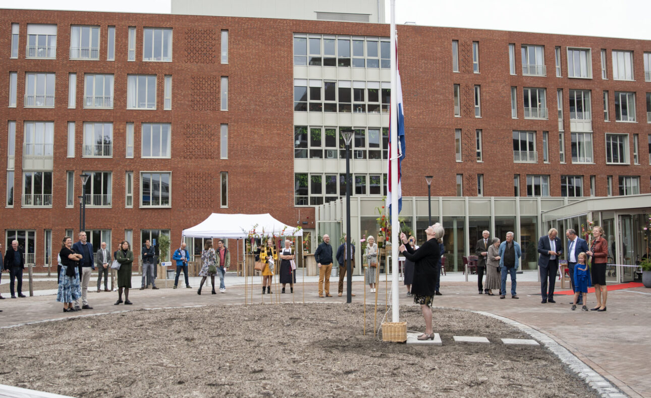 Groep mensen verzameld voor een modern gebouw met rode bakstenen, naast een vlaggenmast en een witte tent op de achtergrond.
