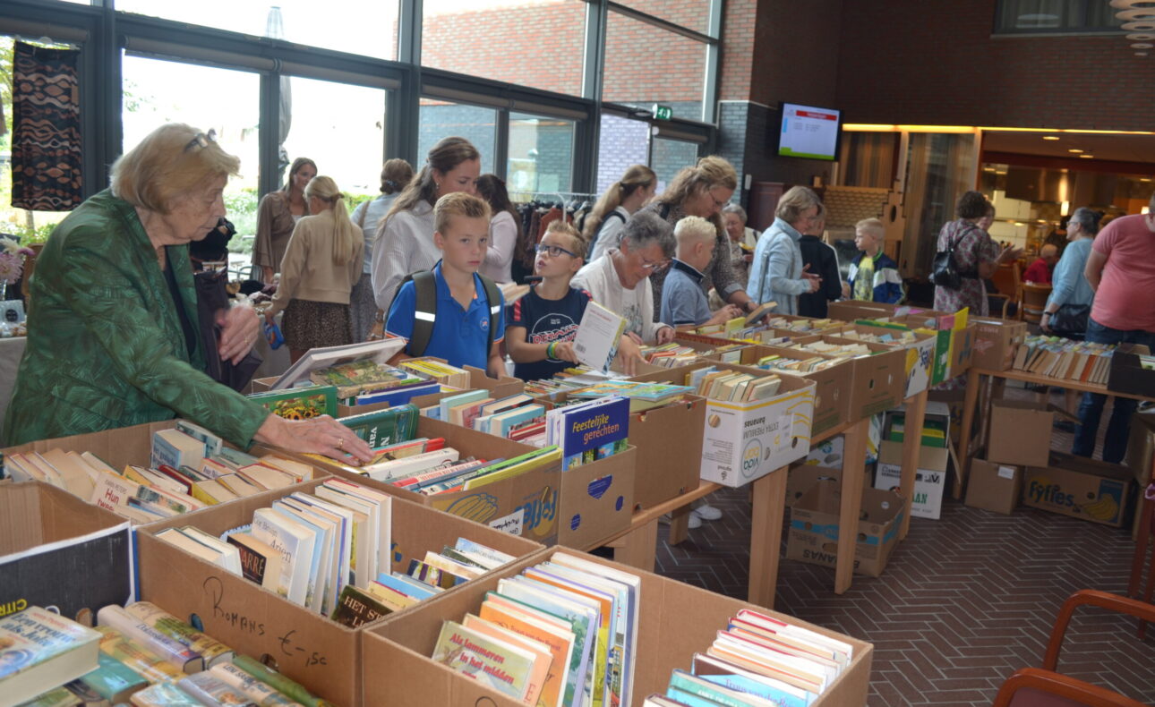 Mensen bladeren door boeken op een boekenmarkt in een ruime, lichte hal. Tafels zijn gevuld met boeken in kartonnen dozen.