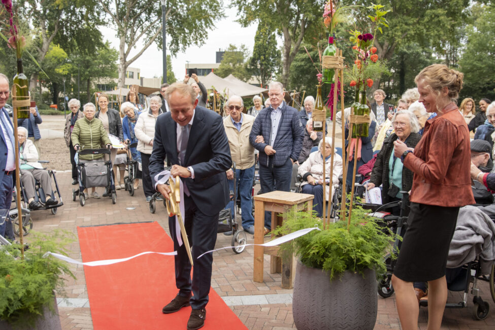 Man in pak knipt lint door op rode loper tijdens een ceremonie, omringd door een groep mensen en planten in potten.