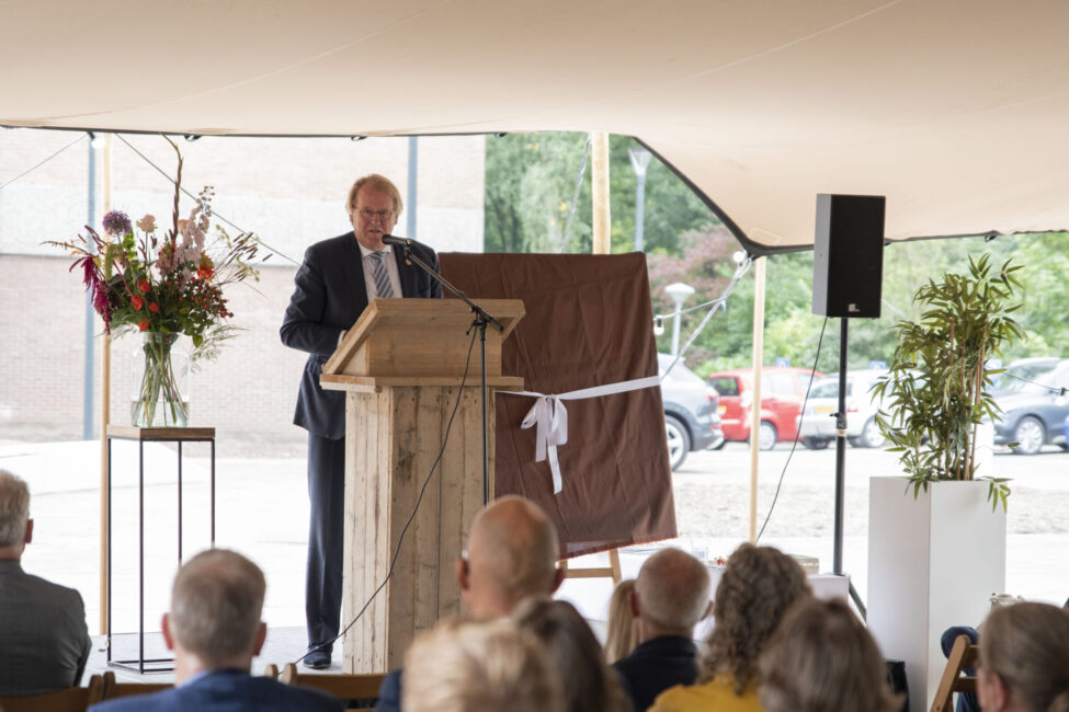 Man spreekt bij een houten lessenaar tijdens een ceremonie. Bloemen en een ingepakt object zijn zichtbaar op de achtergrond.