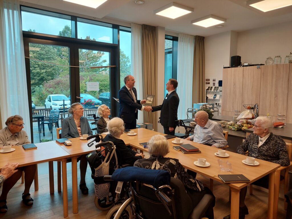 Een groep ouderen zit aan tafels in een lichte ruimte. Twee mannen staan en schudden elkaar de hand. Koffiekopjes op tafel.