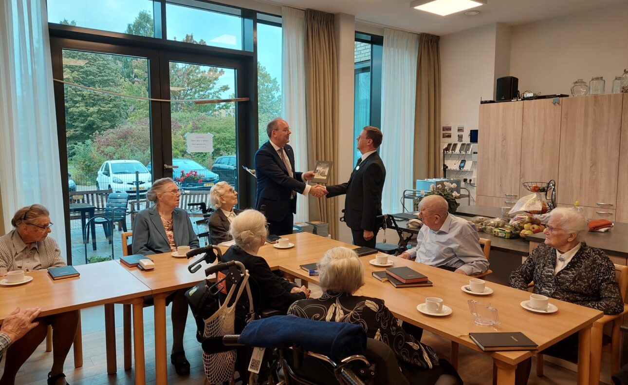 Een groep ouderen zit aan tafels in een lichte ruimte. Twee mannen staan en schudden elkaar de hand. Koffiekopjes op tafel.
