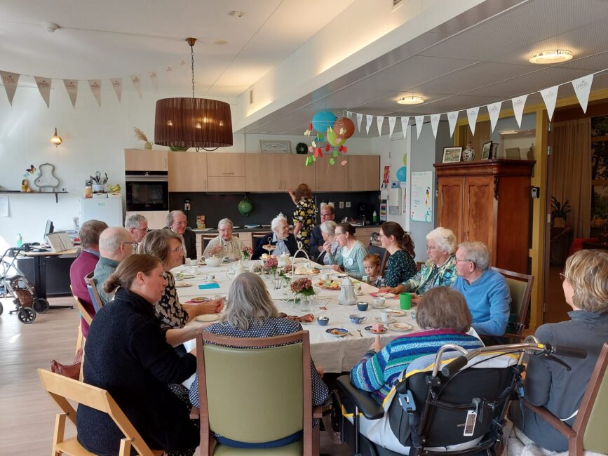 Groep mensen zit rond een tafel voor een high tea in een gezellige ruimte met feestelijke decoraties en slingers.