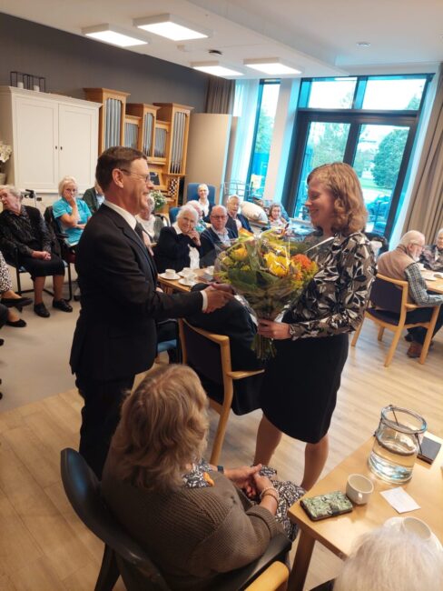 Een vrouw ontvangt een boeket bloemen van een man in een verzorgingstehuis, omringd door zittende ouderen in een zaal.