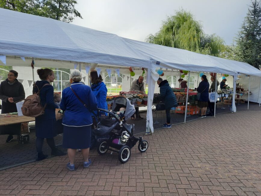 Een markt met mensen die langs kraampjes lopen onder een witte tent. Er is een kinderwagen en diverse producten te zien.
