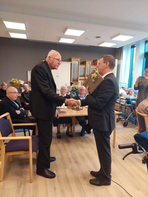 Twee mannen schudden elkaar de hand in een zaal met publiek. Op de achtergrond zitten mensen en staan bloemen.