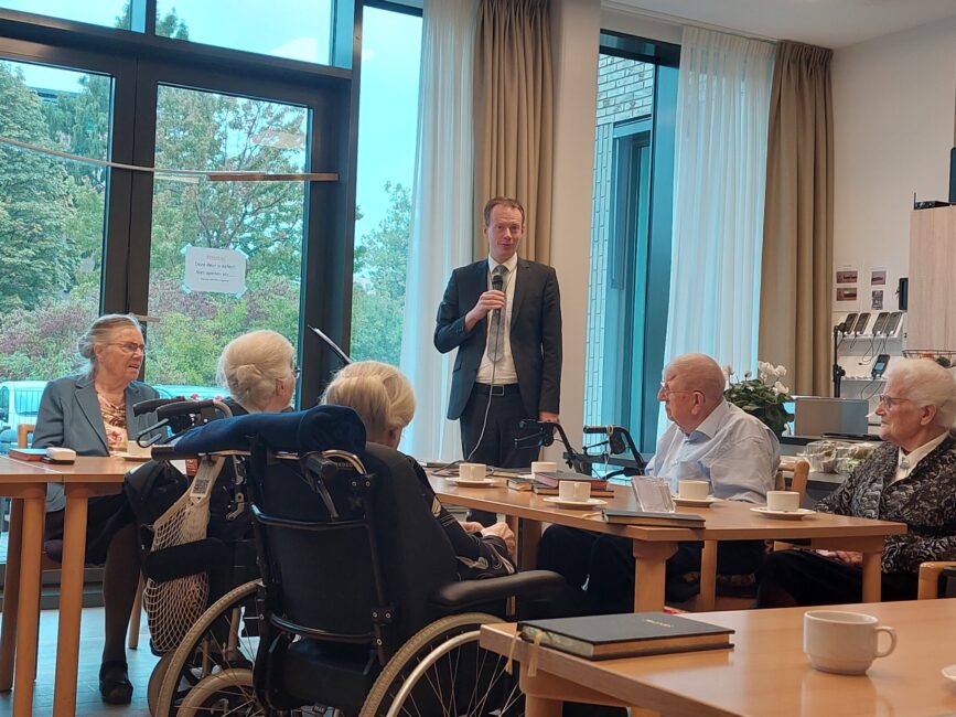 Een man spreekt met een microfoon voor een groep ouderen in een gemeenschappelijke ruimte met grote ramen en tafels.