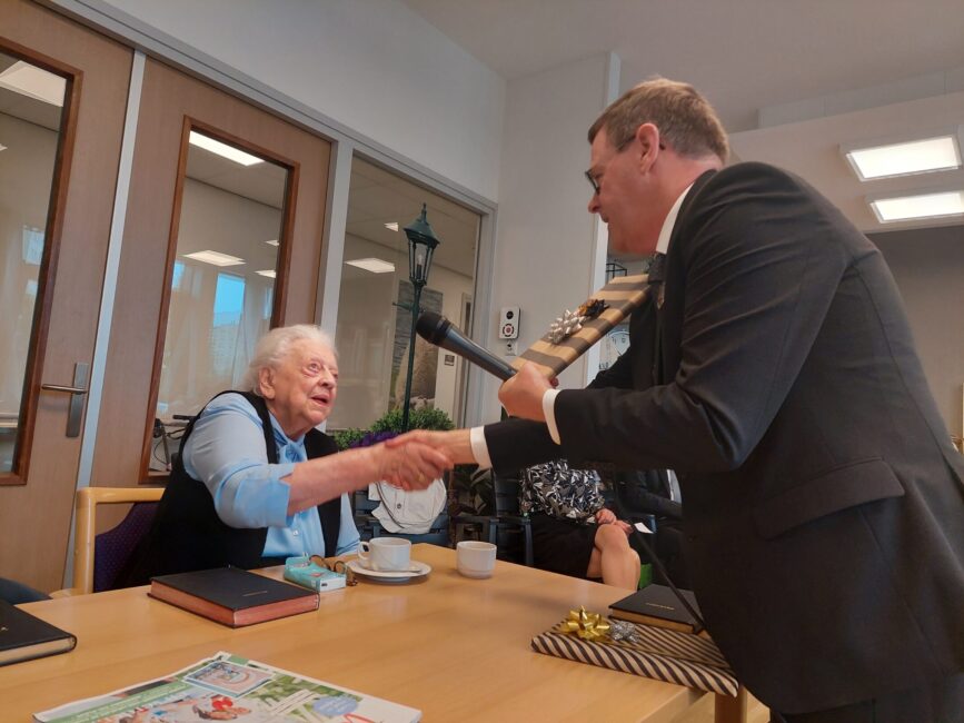 Een oudere vrouw schudt de hand van een man in pak in een vergaderruimte. Op tafel staan kopjes en een krant.