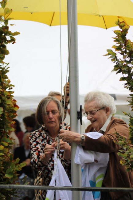 Twee vrouwen hijsen een vlag tijdens een jubileumweek. Ze staan onder een gele paraplu, omringd door groene planten.