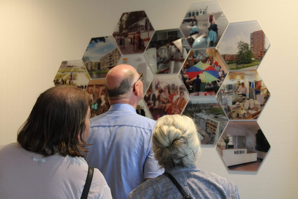 Drie mensen bekijken een fotowand met zeshoekige foto's die verschillende scènes en gebouwen tonen in een expositieruimte.